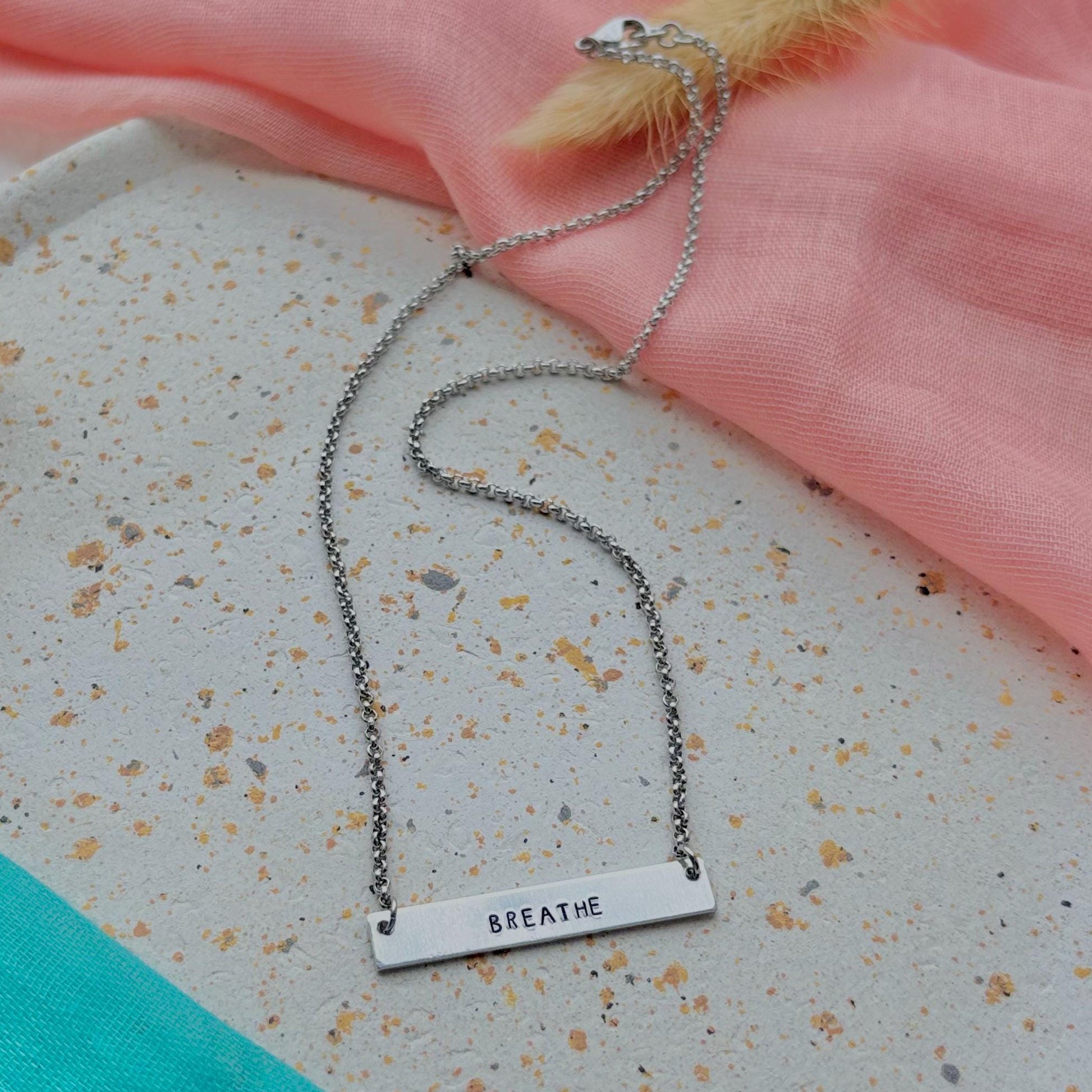 Necklace with a bar pendant reading 'BREATHE' on a textured surface with pink fabric and feathers.