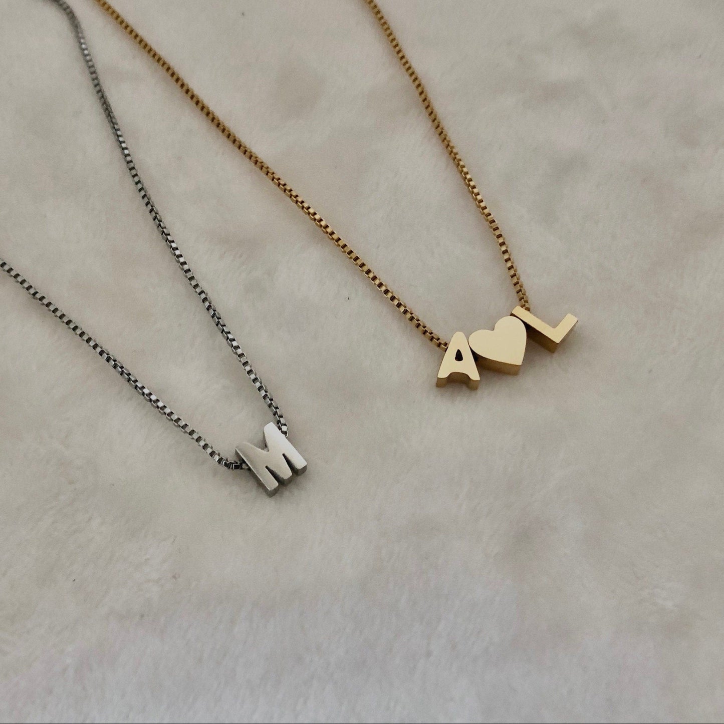 Two necklaces on a beige background, one gold with 'M' and 'L' charms, the other silver with 'M' charm.