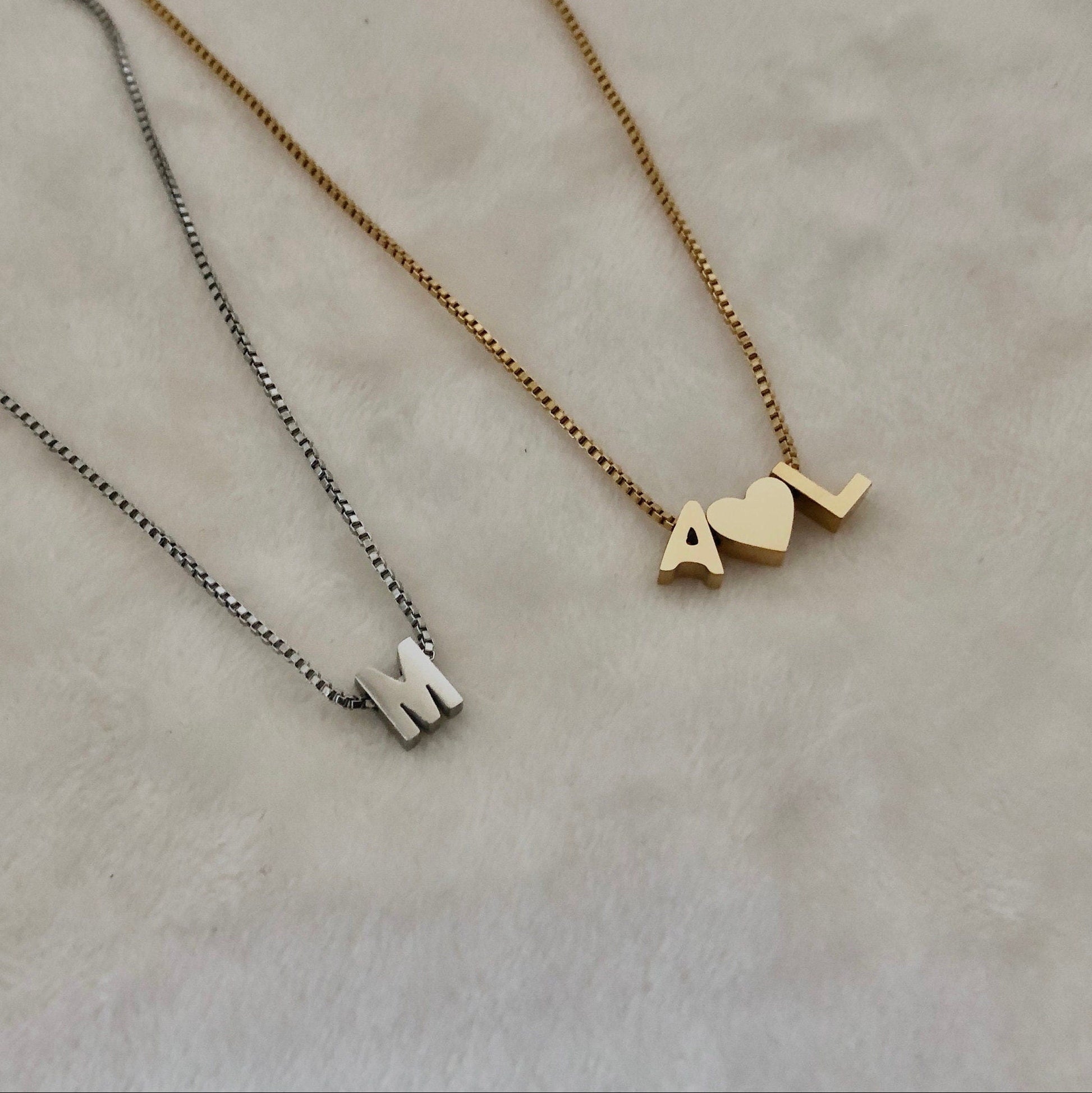 Two necklaces on a beige background, one gold with 'M' and 'L' charms, the other silver with 'M' charm.