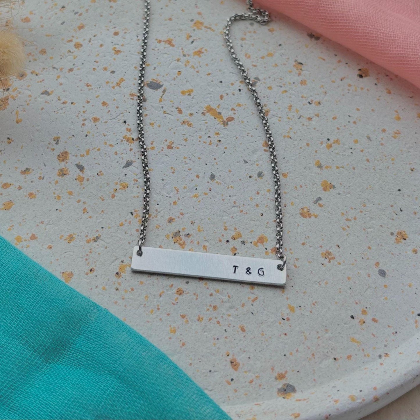 Silver bar necklace with 'T & G' engraving on a textured surface