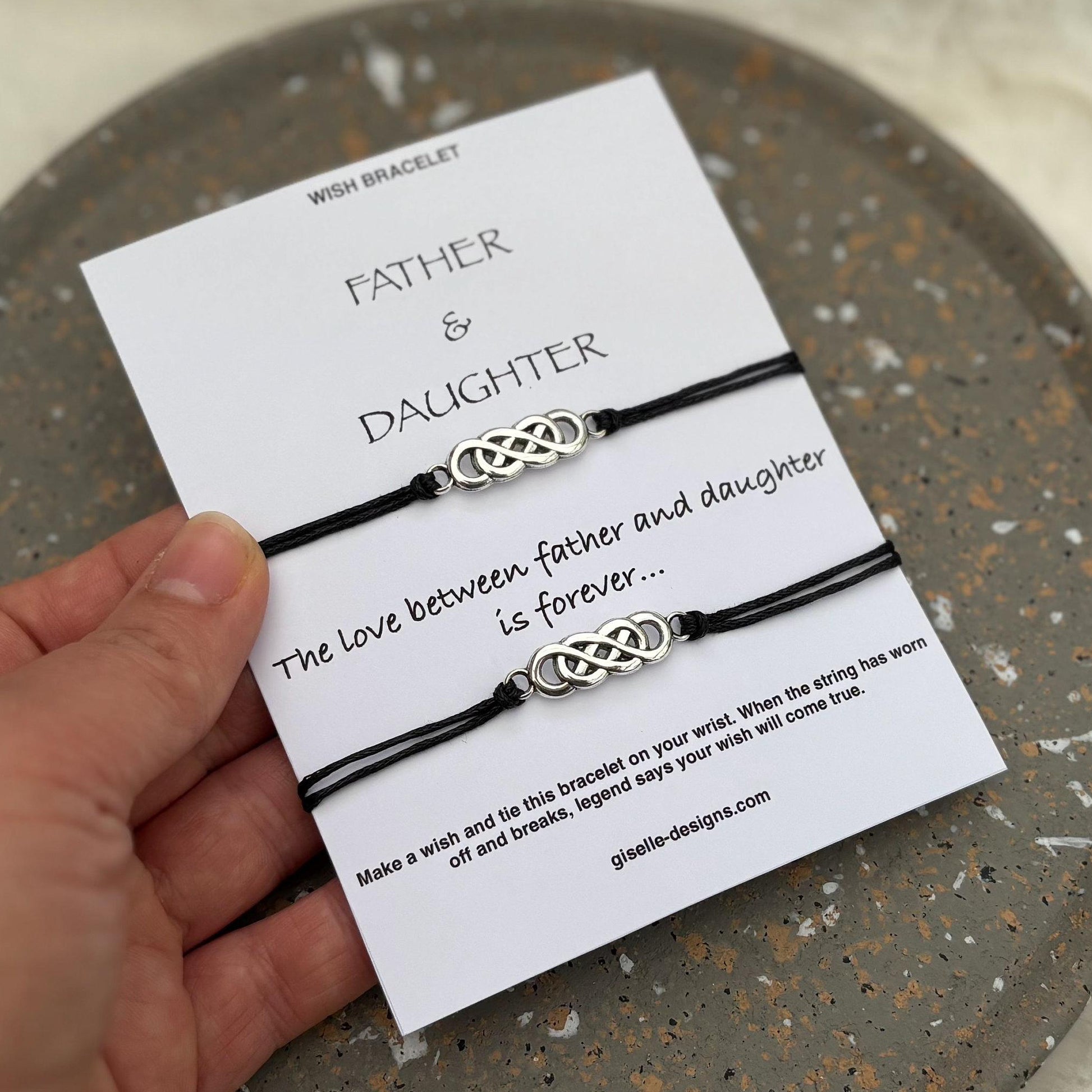 Two black braided bracelets with silver Celtic knot designs on a card labeled 'Father & Daughter' held by a hand.
