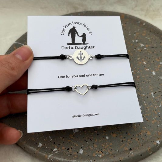 Two black cord bracelets with heart and anchor charms on a card, held by a hand.