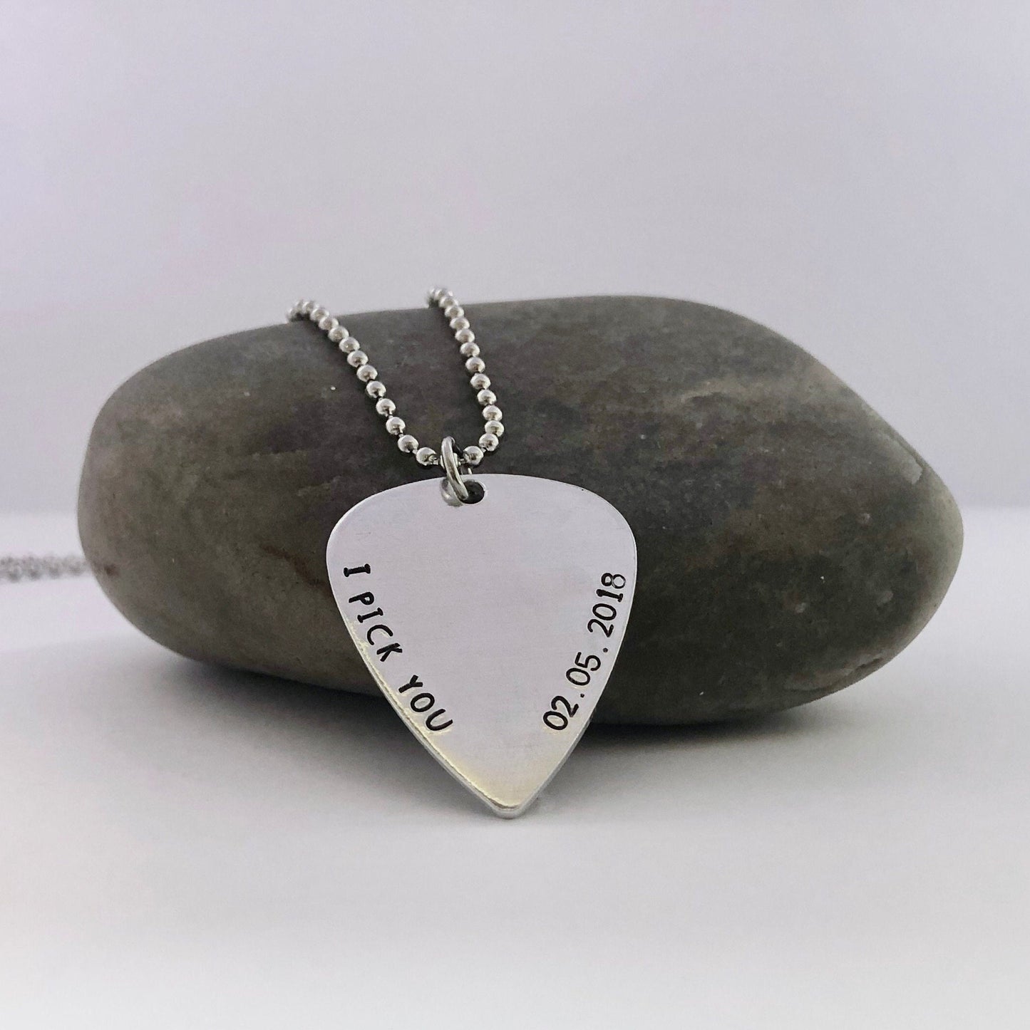 Silver guitar pick-shaped pendant with 'I Pick You' and date on a stone background