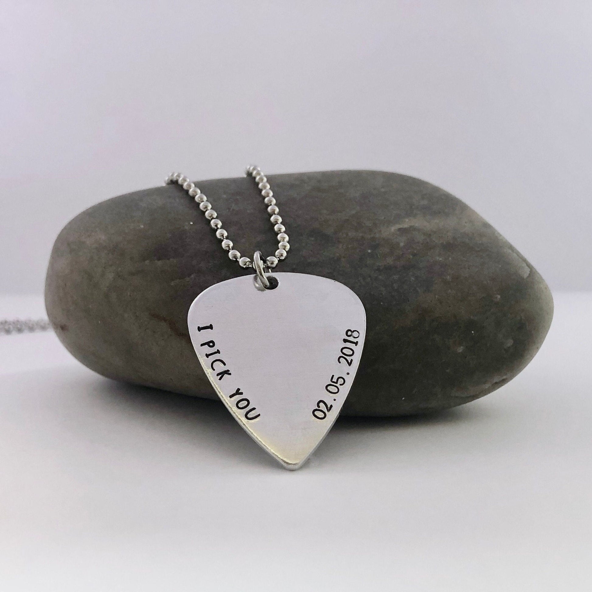 Silver guitar pick-shaped pendant with 'I Pick You' and date on a stone background