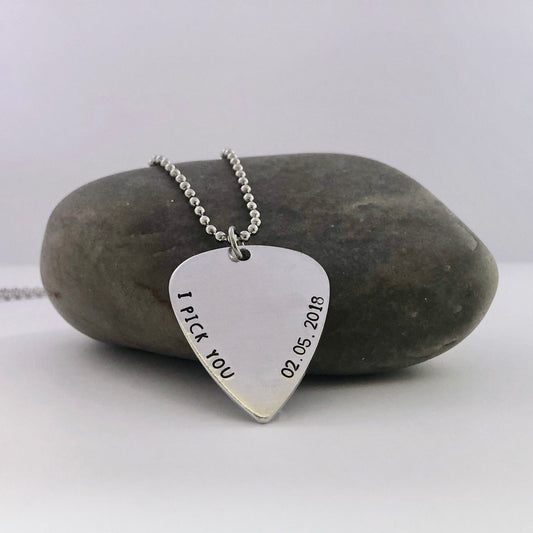 Silver guitar pick-shaped pendant with 'I Pick You' and date on a stone background