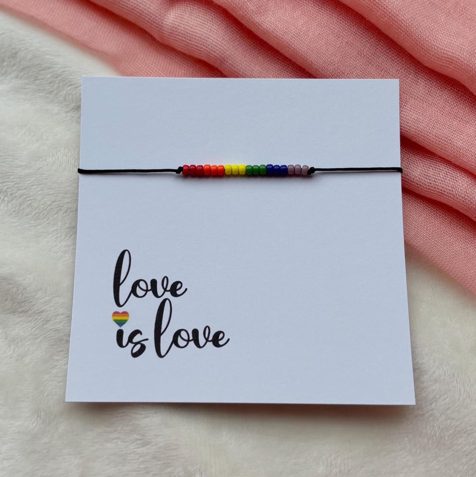 Bracelet with rainbow beads on a card labeled 'love is love' against a pink fabric background