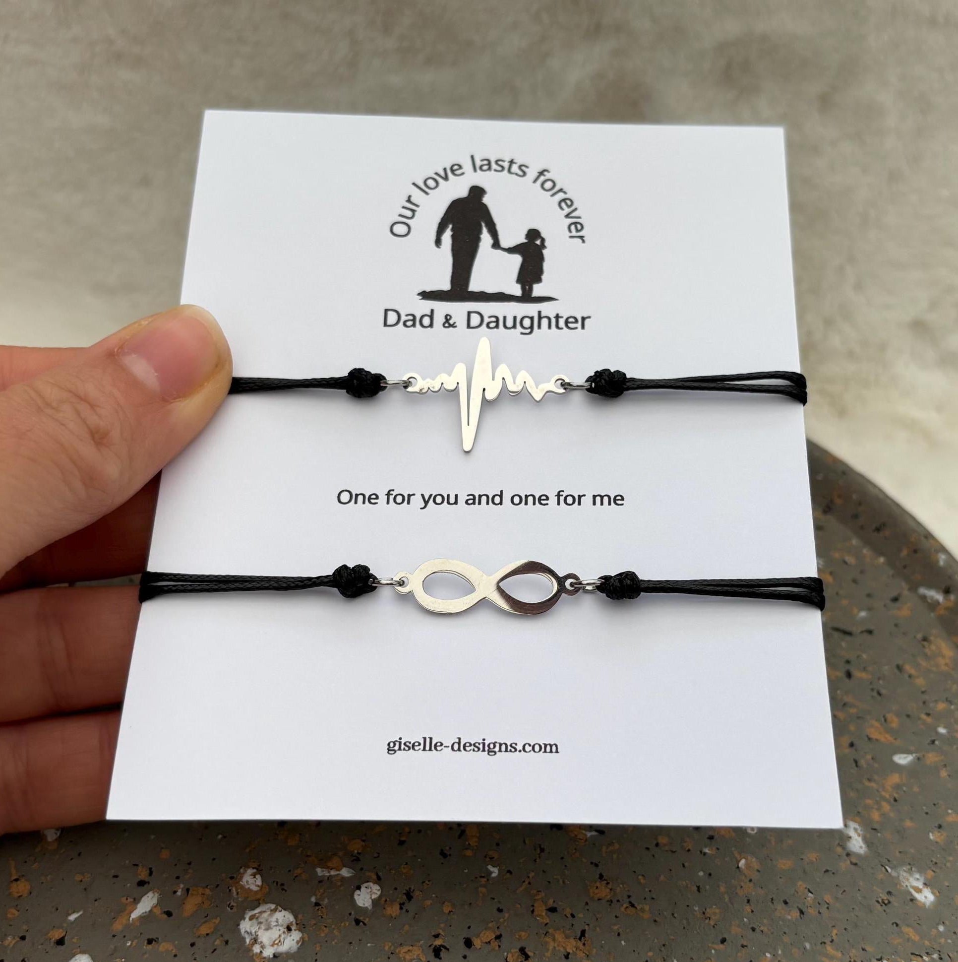 Two black bracelets with gold charms on a card held by a hand, with a neutral background.