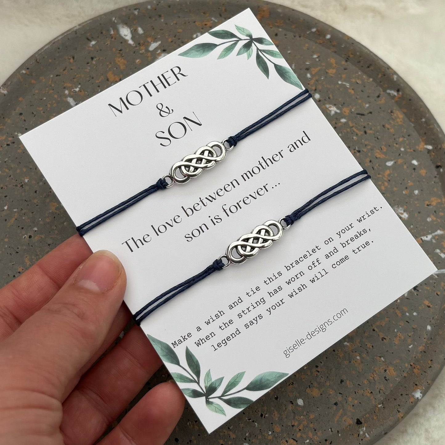Two braided bracelets on a card with 'Mother & Son' text, held by a hand on a textured surface.
