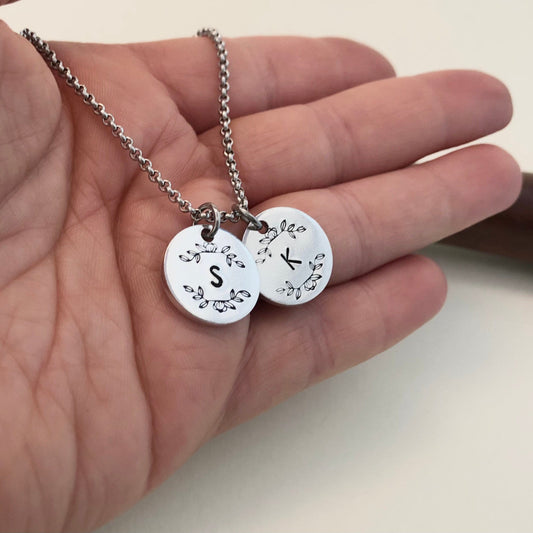 Two silver pendants with initials held in a hand against a neutral background
