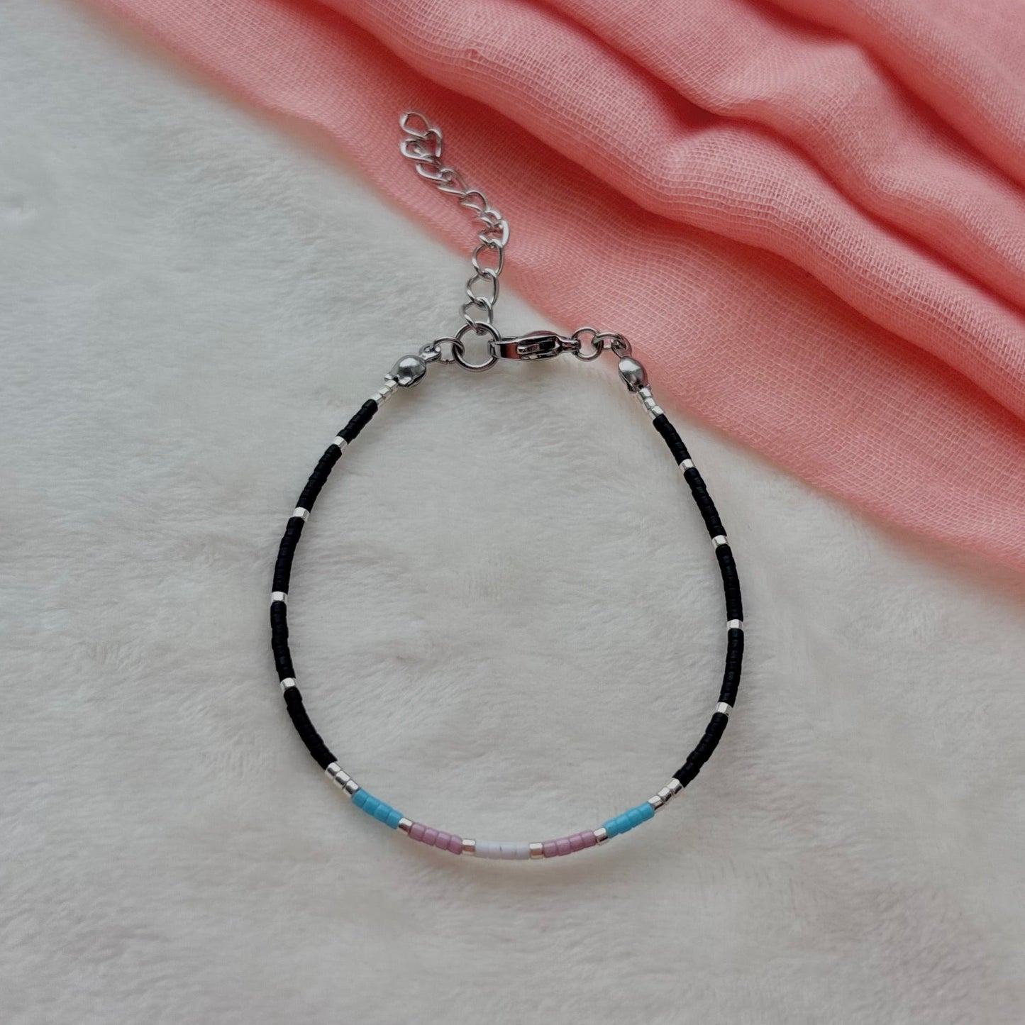 A delicate beaded bracelet with black and white beads, accented by small blue and pink beads, resting on a soft white textured surface with a draped pink fabric in the background.