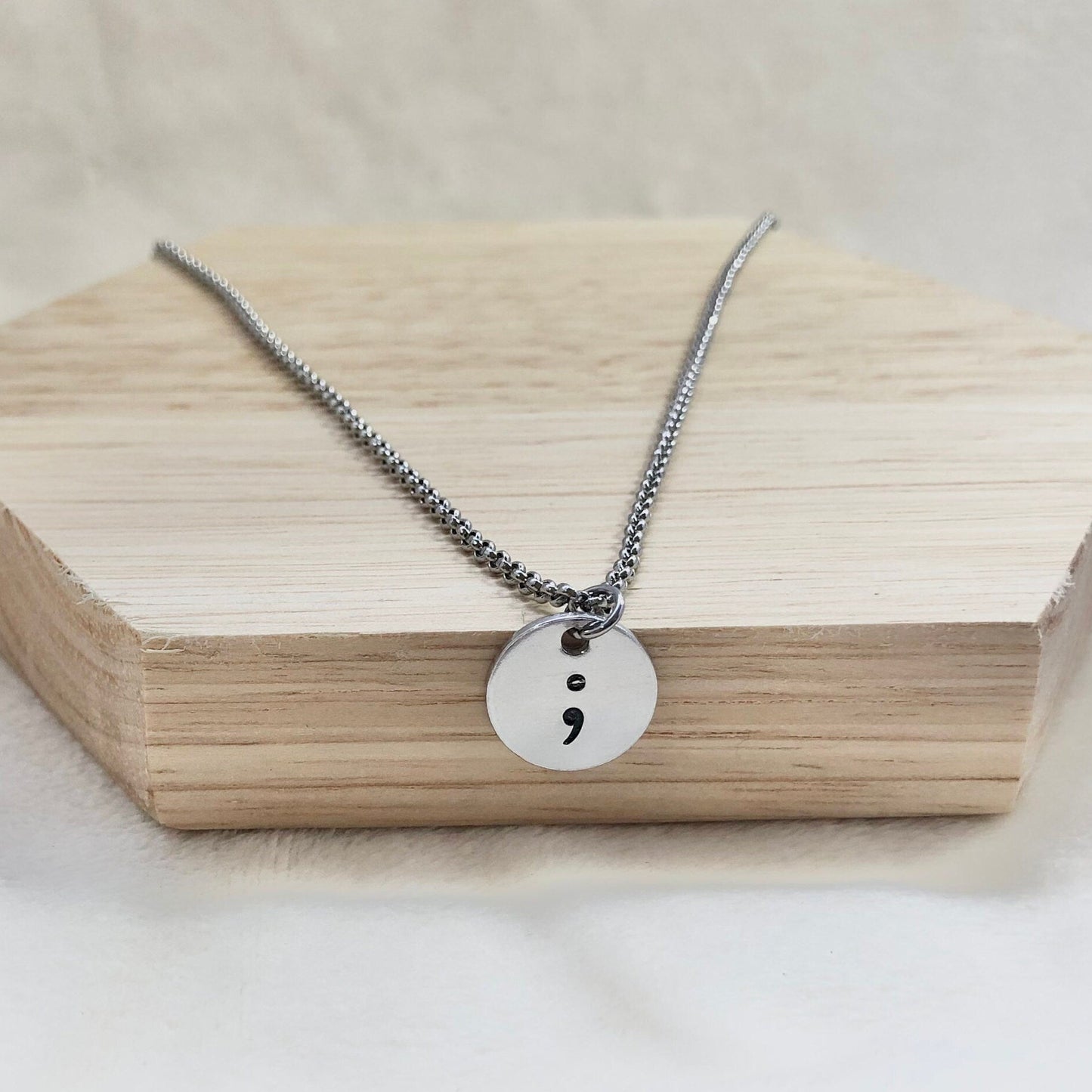 Necklace with a round pendant featuring a semicolon on a wooden block.