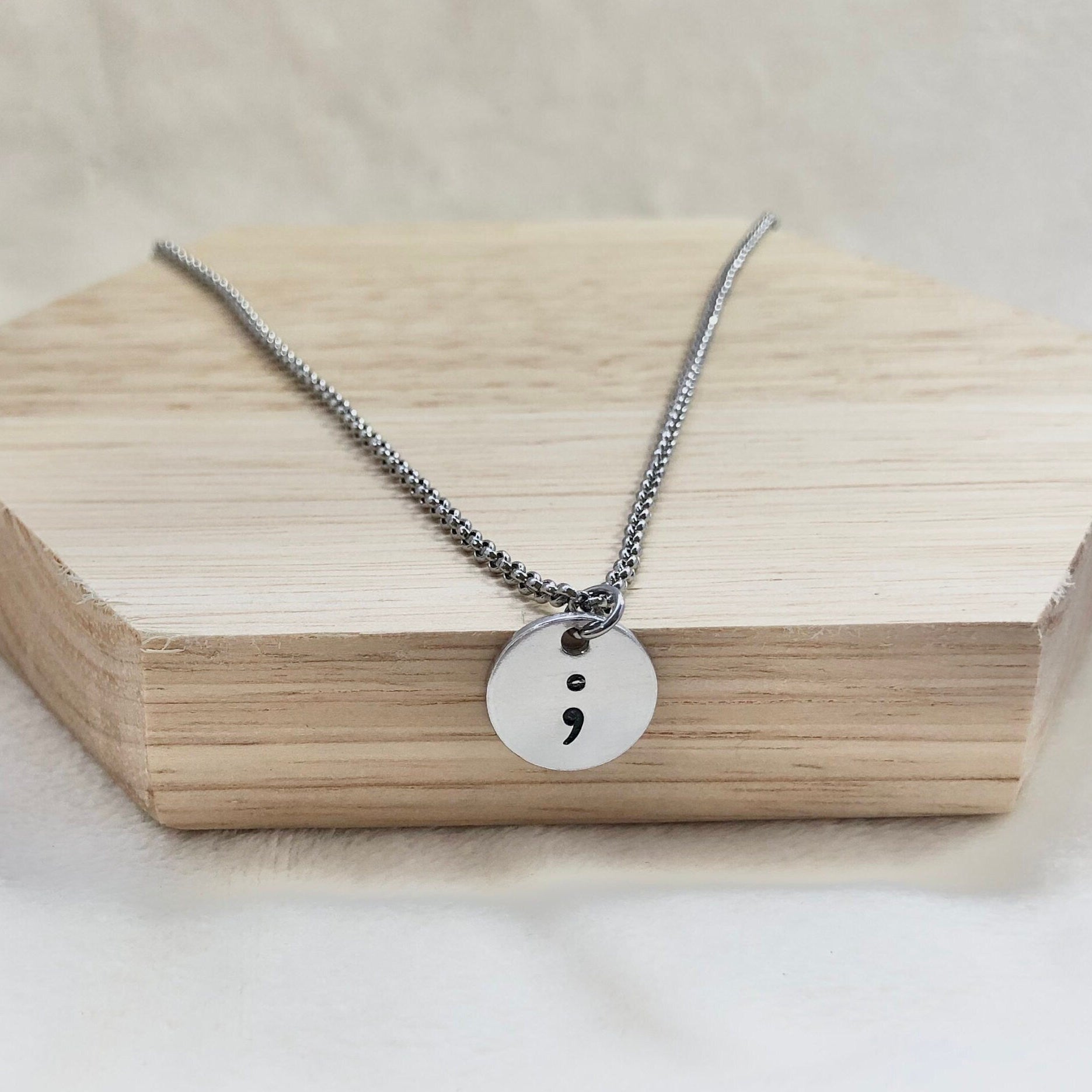Necklace with a round pendant featuring a semicolon on a wooden block.