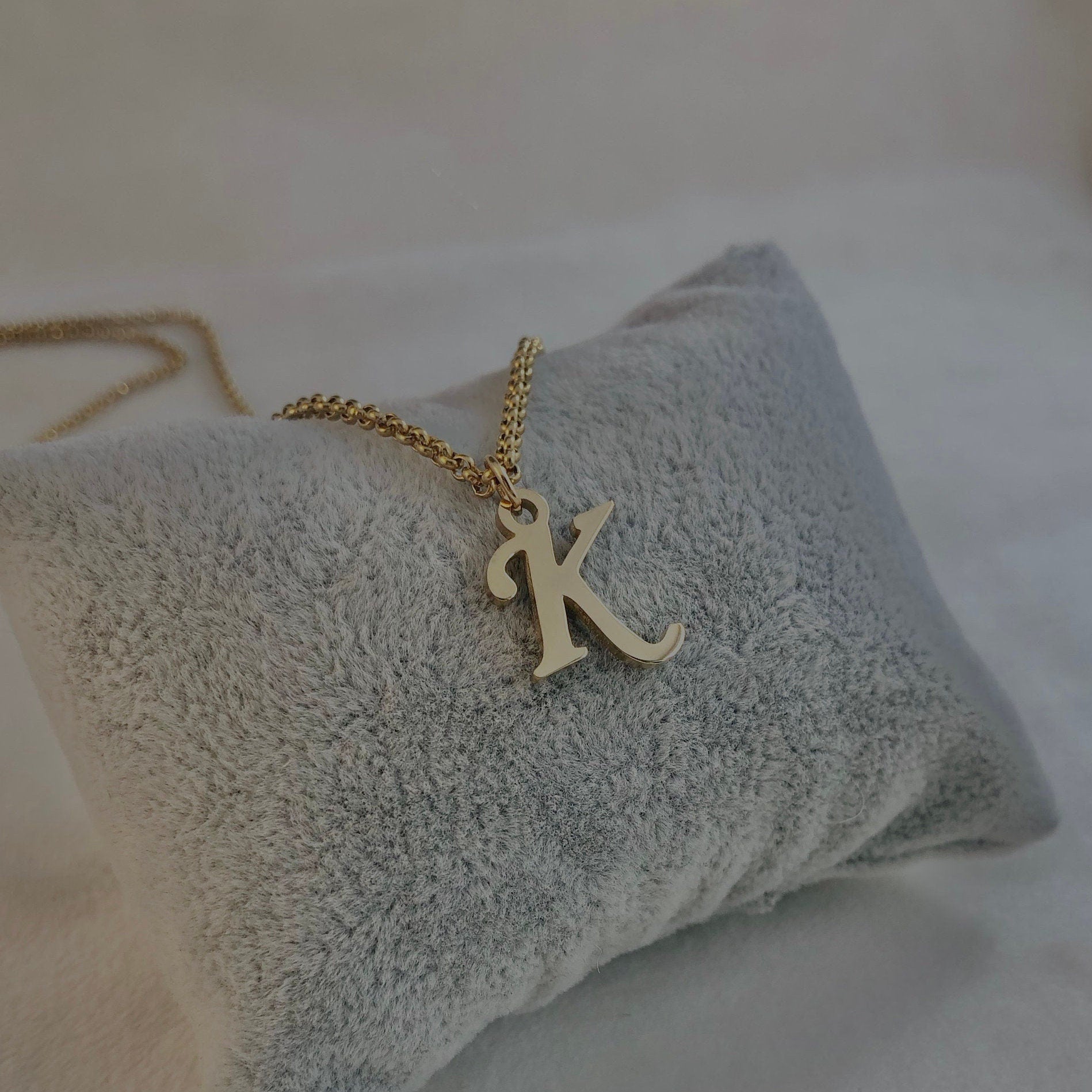 Gold necklace with a 'K' pendant on a textured gray pillow.