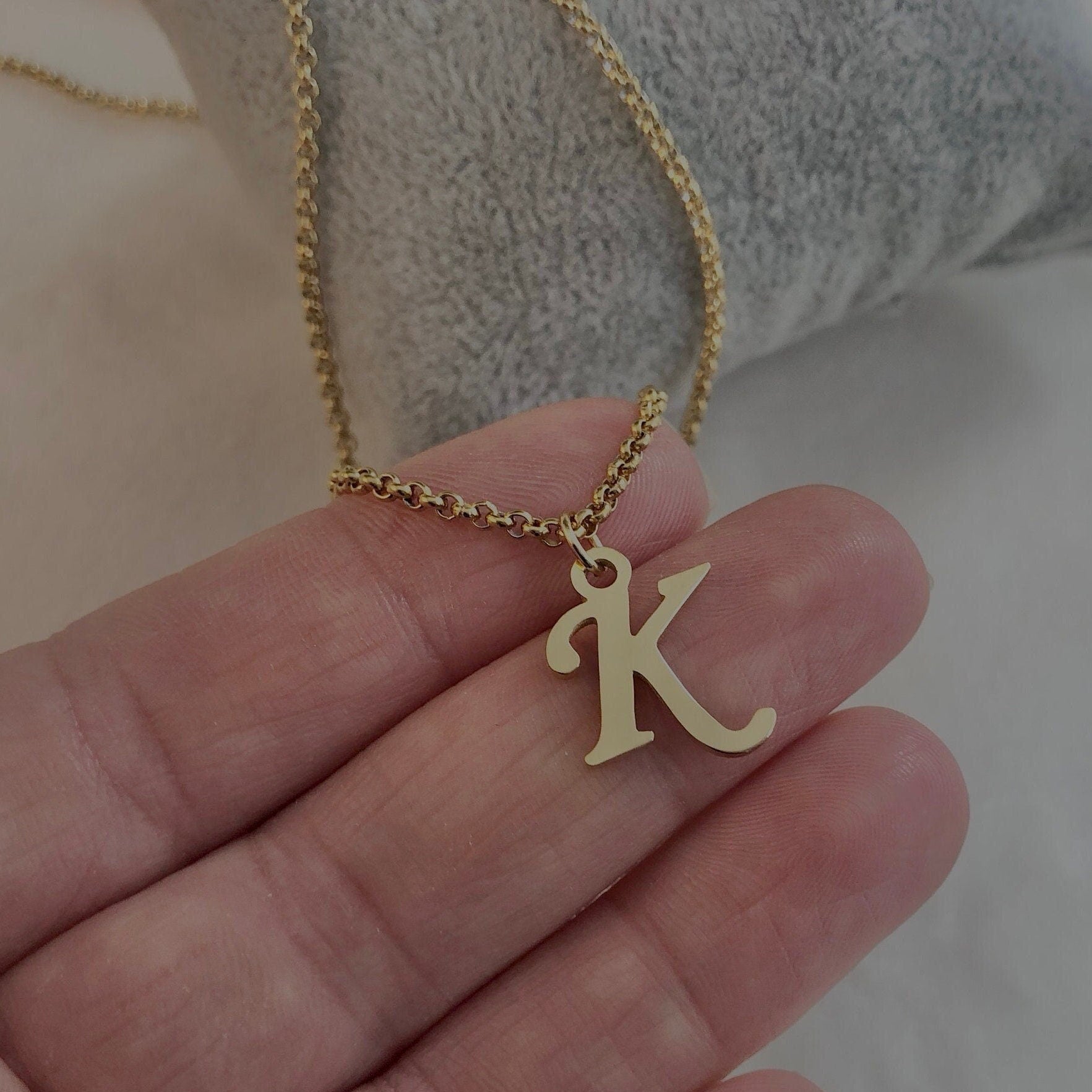 Gold necklace with a 'K' pendant held in a hand against a neutral background