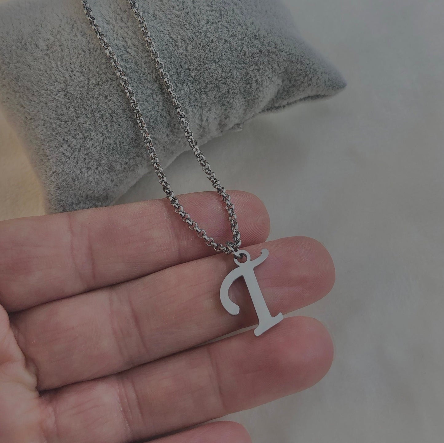 Hand holding a silver necklace with a 'T' pendant against a neutral background