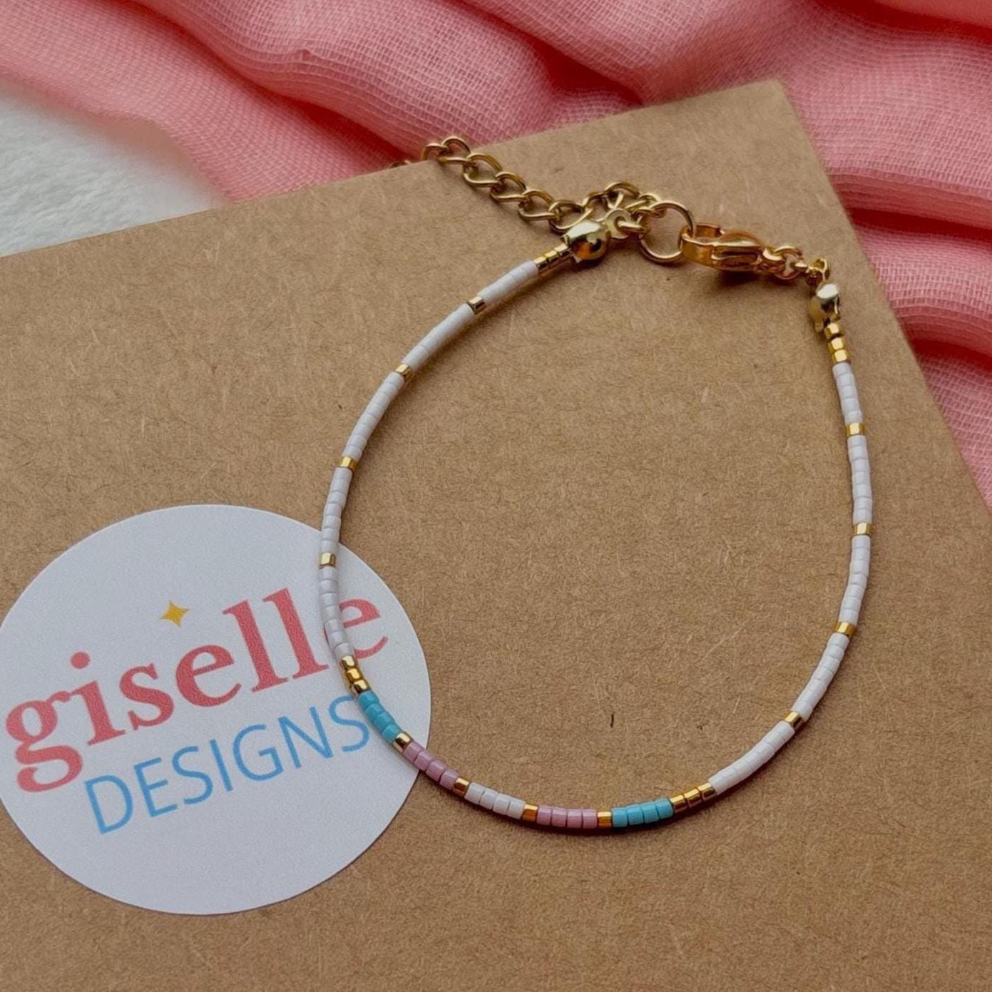 Bracelet with multicolored beads on a brown card with 'giselle DESIGNS' label, pink fabric background