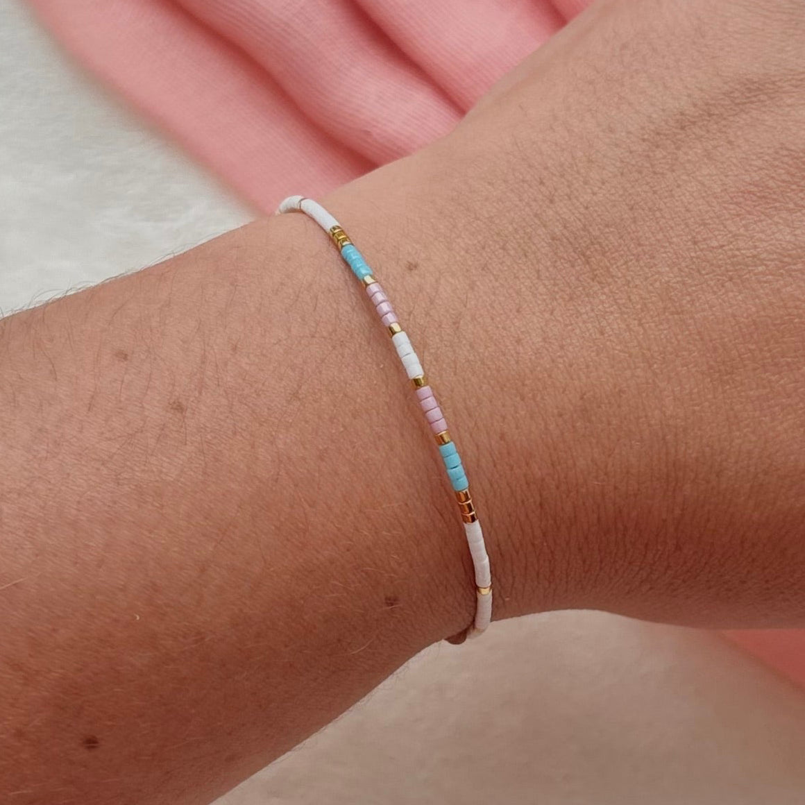 A delicate beaded bracelet with white, light blue, and light pink beads, shown worn on a person's wrist.