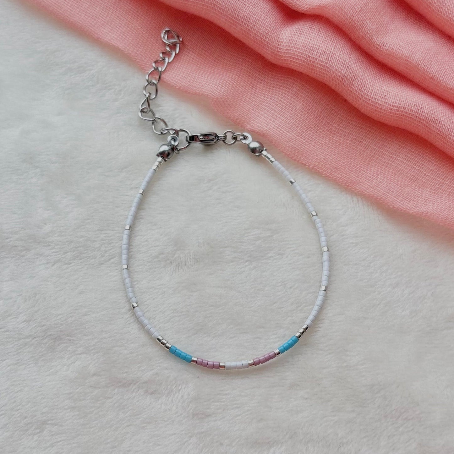 A delicate silver-toned beaded bracelet with small blue, pink, and white beads, resting on a soft white textured surface next to a draped pink fabric.