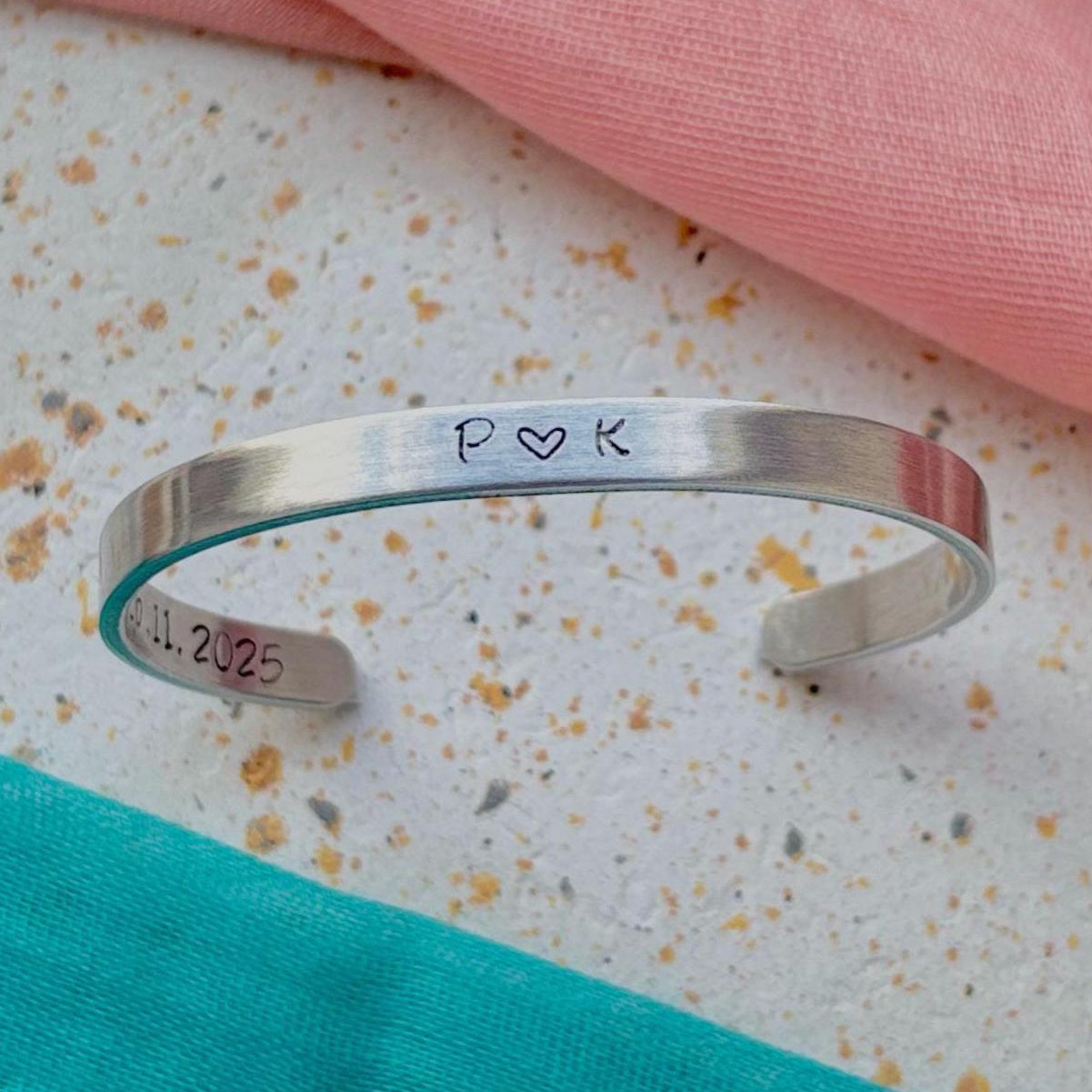 Silver bracelet with engraved initials and date on a textured surface with pink and green fabric.