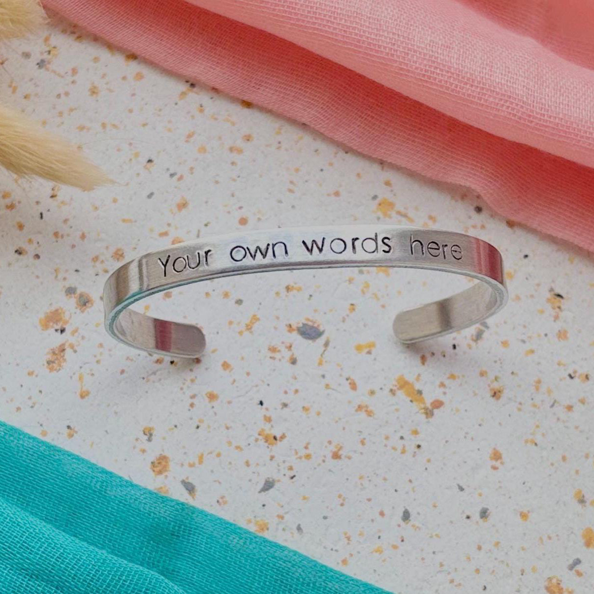 Silver adjustable bracelet with 'Your own words here' engraving on a textured surface with pink and green fabric.