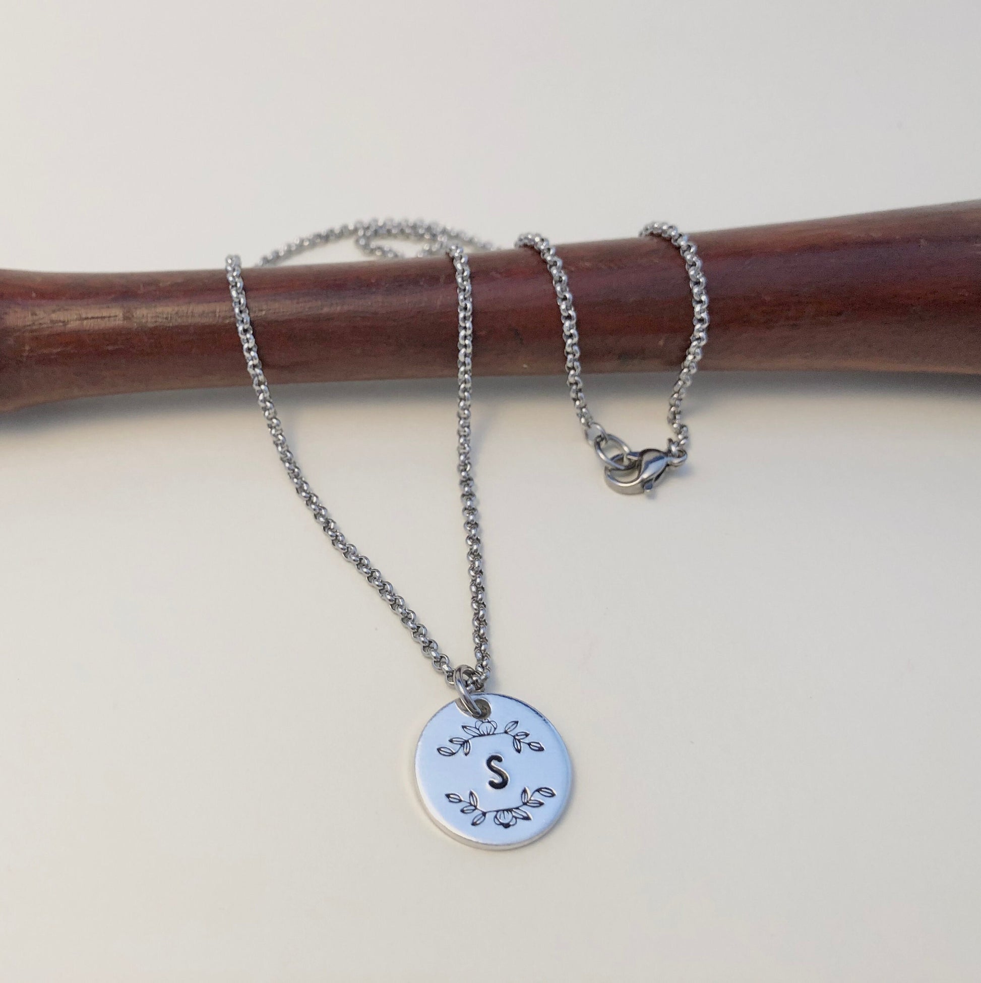 Silver necklace with a round pendant featuring the letter 'S' on a wooden stick.