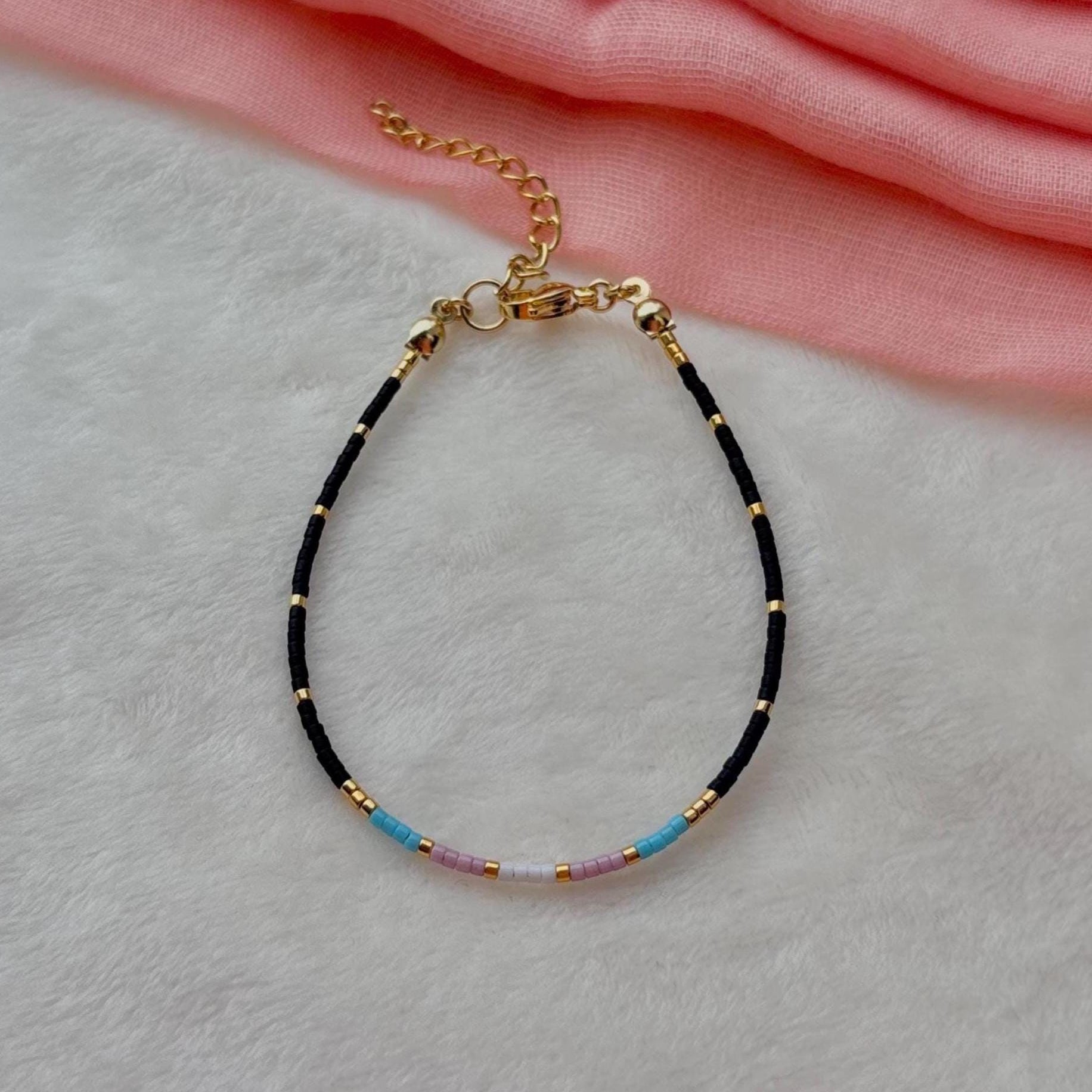 Trans flag bracelet with black and gold beads on a light gray surface with pink fabric in the background