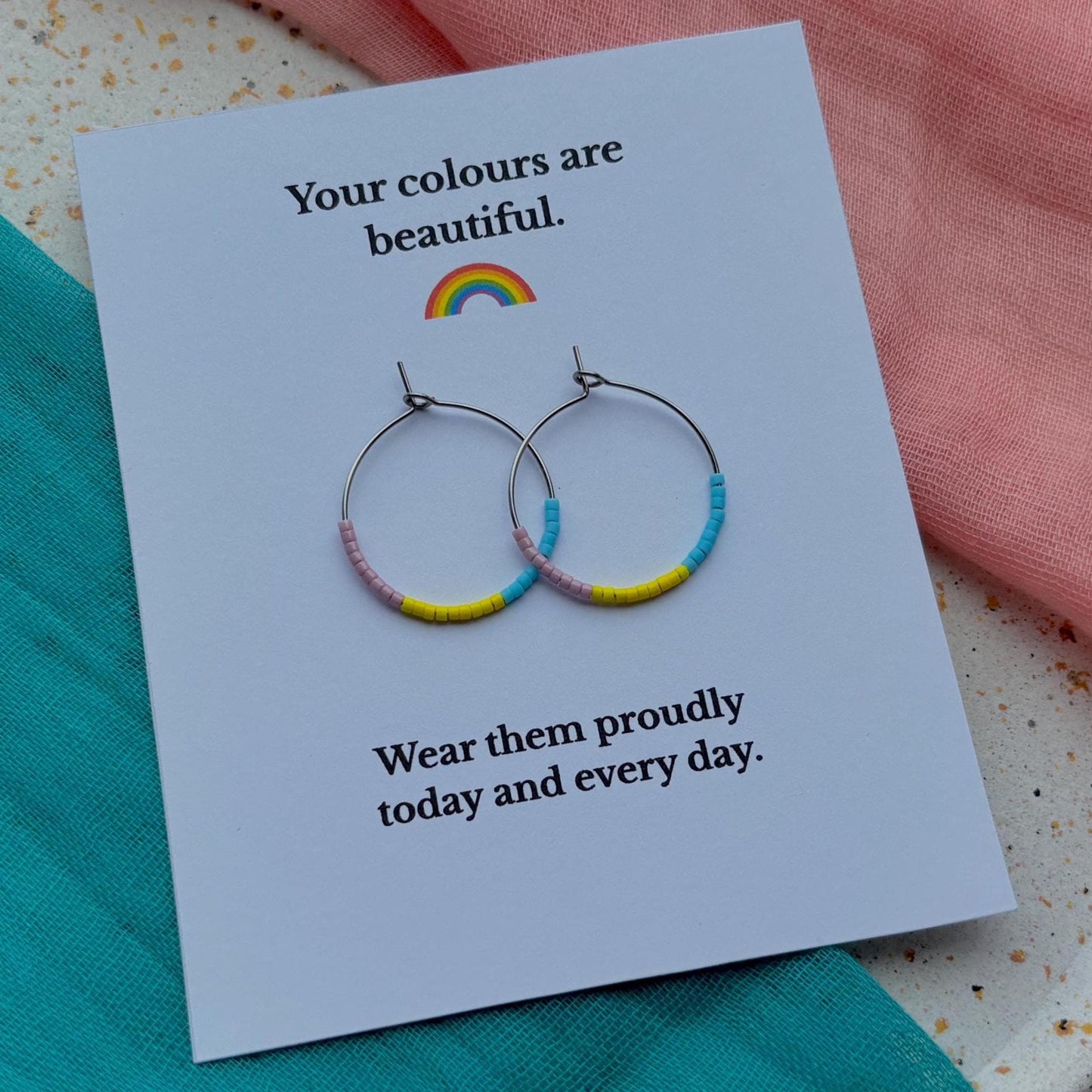 Earrings with colorful bands on a card with text and a rainbow, placed on a textured surface.