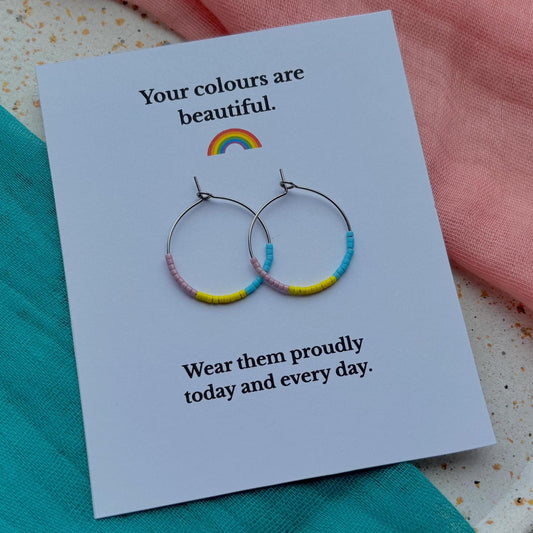Earrings with colorful bands on a card with text and a rainbow, placed on a textured surface.
