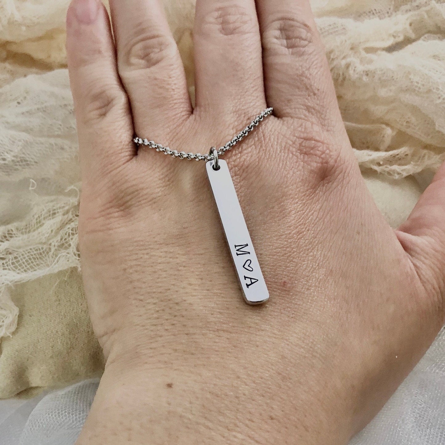 Hand wearing a silver necklace with a bar pendant on a textured fabric background