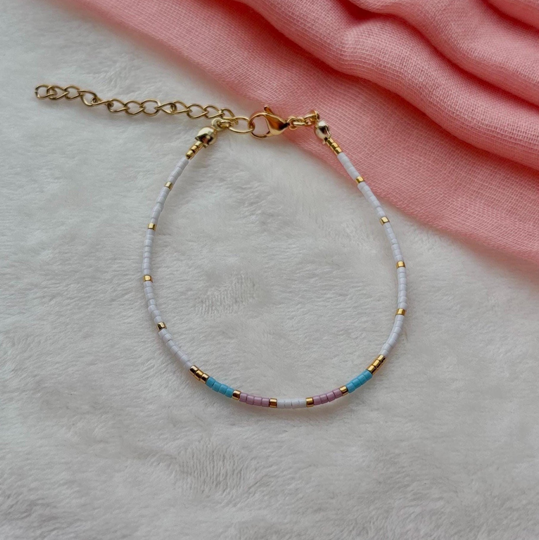 Bracelet with trans flag beads on a light gray surface with pink fabric in the background