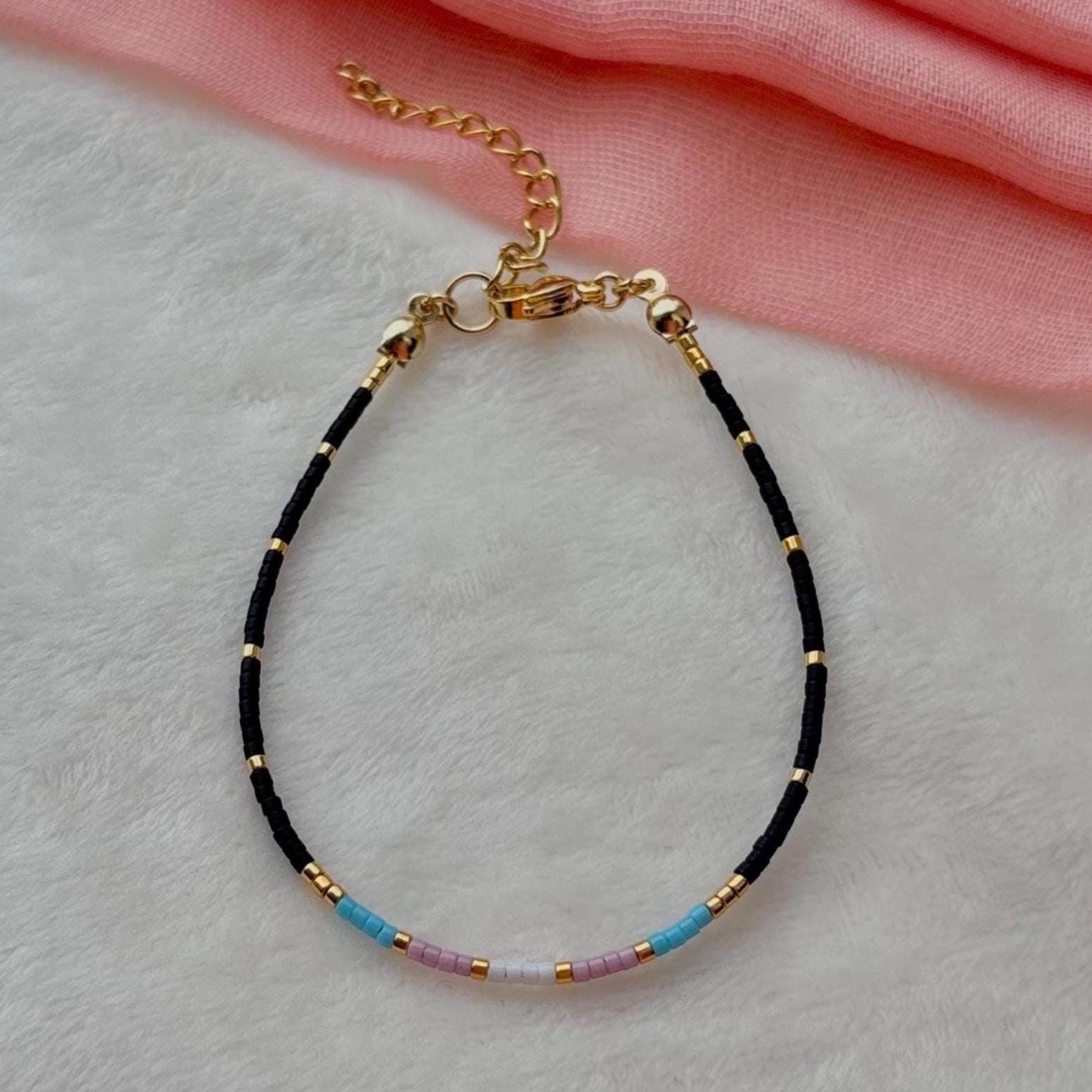 Trans flag bracelet with black and gold beads on a light gray surface with pink fabric in the background
